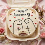 A round white cake in a beige take-out container lid, decorated with 'Happy 1st Anniversary' in black icing, small red heart candies, two simple black line figures, and a small 'Galaave' logo at the bottom. The cake is styled on pink satin fabric with roses, candlesticks, and forks.