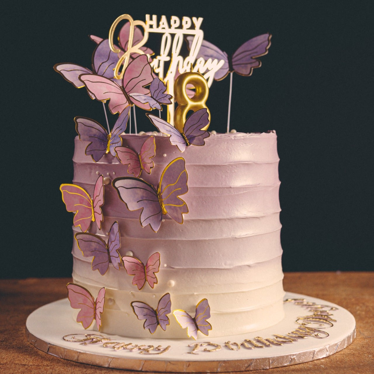 A beautifully decorated cake with multiple layers, covered in pink and white frosting, adorned with purple and gold butterflies, and a 'Happy Birthday' topper.