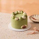 A round green cake with white piped cream decorations on top, sitting on a white plate on a speckled beige table. A small oval cake topper with the brand name 'Cateraux' is attached to the front right side.