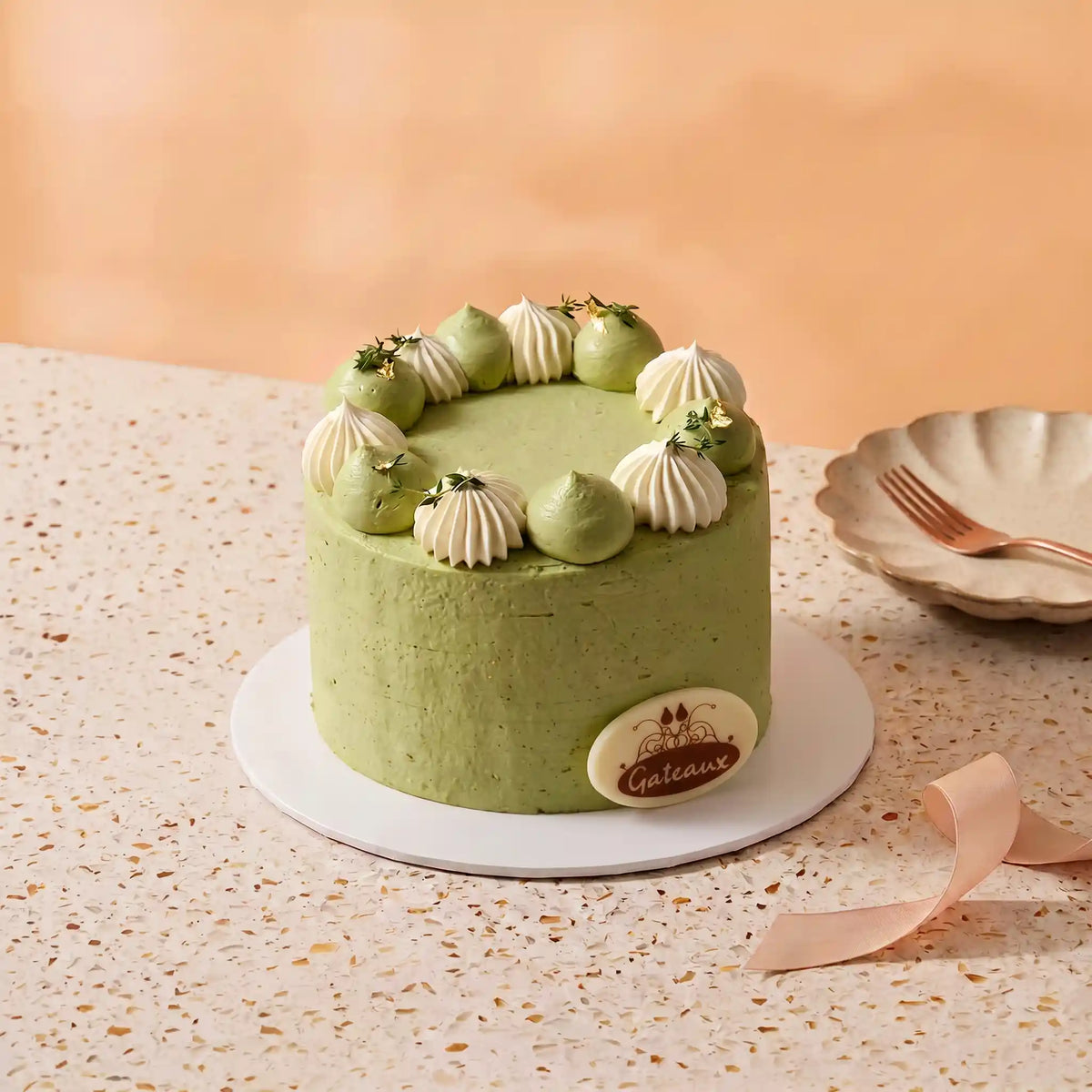 A round green cake with white piped cream decorations on top, sitting on a white plate on a speckled beige table. A small oval cake topper with the brand name 'Cateraux' is attached to the front right side.