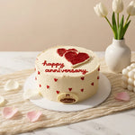 A round anniversary cake with white frosting, red heart decorations, and 'happy anniversary' written in red icing on top. The cake sits on a white plate on a beige textured runner, with pink rose petals scattered around, a white vase of tulips, and a decorative pearl cluster in the background.