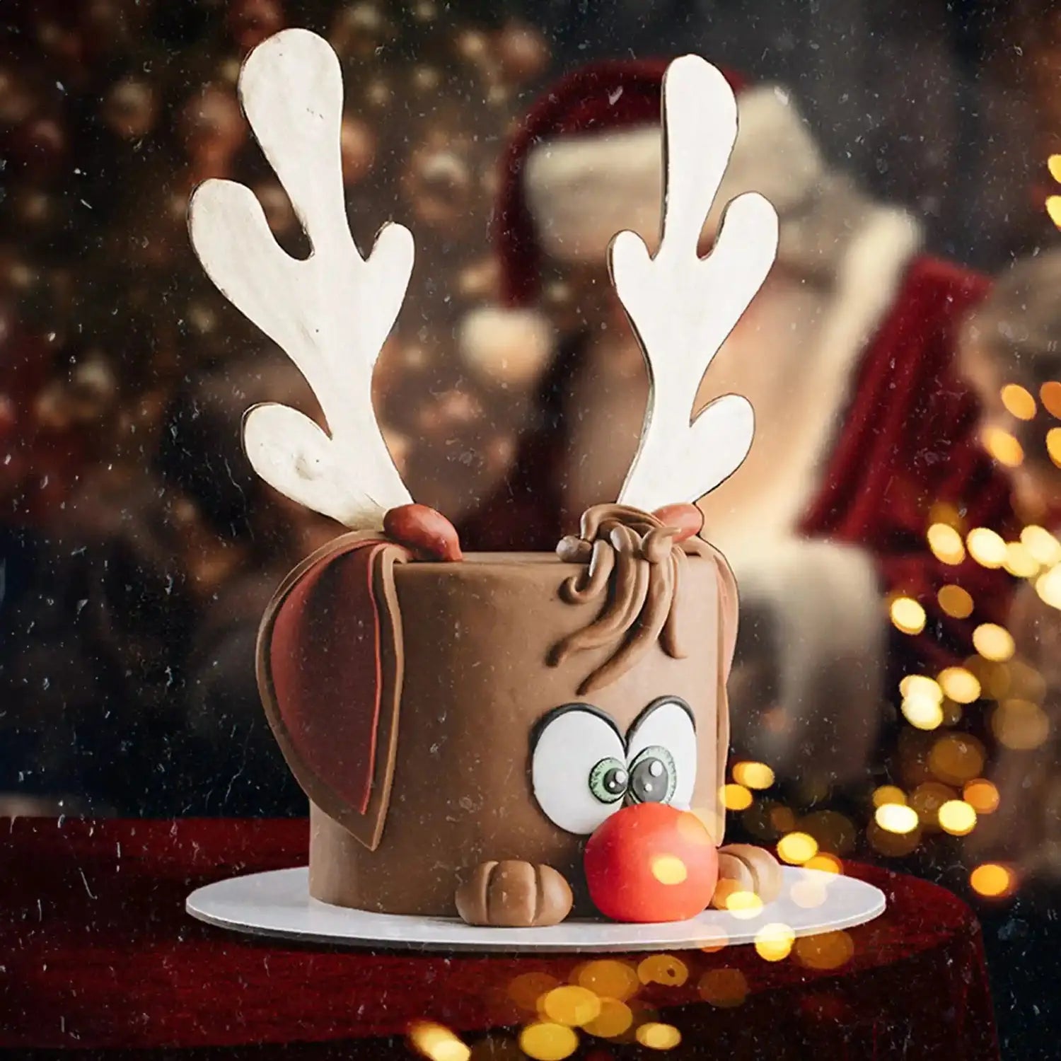 Chocolate reindeer cake with white antlers and a red nose on a blurred festive background.