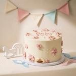 A round white frosted cake decorated with pink flower motifs and small pink heart details on top, displayed on a white round stand. The cake is styled with a white wooden rainbow and rocking horse decoration, pastel ribbons, and a pastel bunting banner in the background.