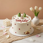 A white frosted birthday cake with red heart decorations and 'happy 2 years anniversary' written on top, decorated with two small green frog figurines. The cake sits on a marble surface with a beige textured runner, surrounded by tulips in a white vase and a decorative candle against a warm beige background.