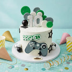 A decorated birthday cake with a white frosting base, green and gray polka dots, a large white number 10 topper, the word 'BASHED' in green outline letters, and a gray video game controller decoration. The cake sits on a white round board with party cups, confetti, and streamers on a light blue background.