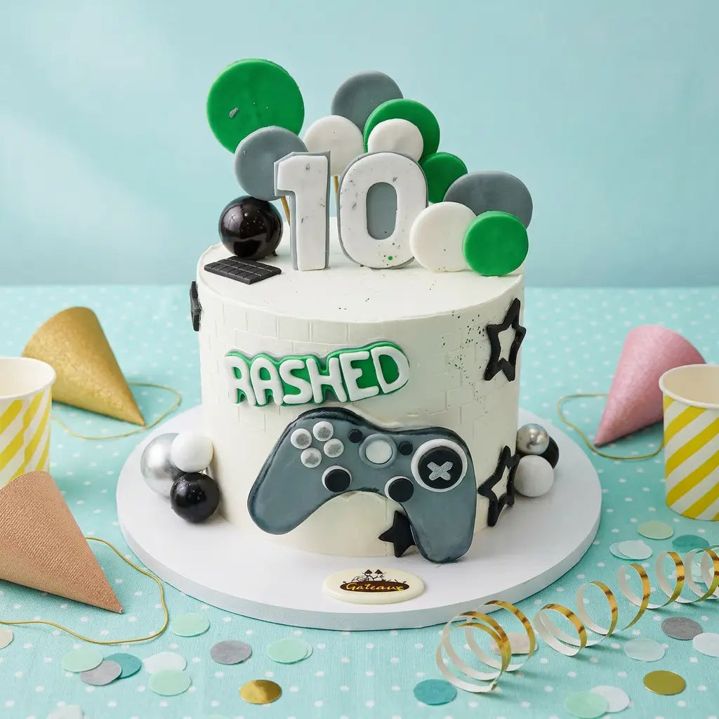 A decorated birthday cake with a white frosting base, green and gray polka dots, a large white number 10 topper, the word 'BASHED' in green outline letters, and a gray video game controller decoration. The cake sits on a white round board with party cups, confetti, and streamers on a light blue background.