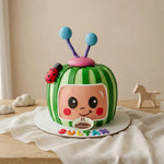 A custom 3D Cocomelon-themed birthday cake shaped like a green striped watermelon with a cartoon face, blue antennae, and a ladybug decoration, displayed on a wooden table with a neutral background.