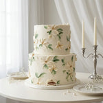 A two-tier white wedding cake decorated with 3D white flowers and green leaves, displayed on a round white table with a silver candlestick and white drapery in the background.