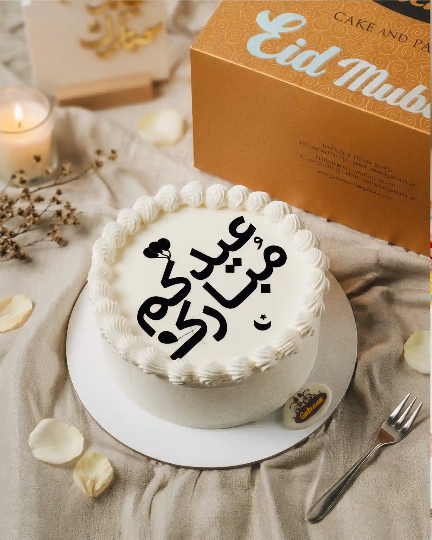 White cake with Arabic text on a beige surface with an 'Eid Mubarak' box in the background.