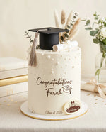 Graduation-themed cake with a black cap and diploma, decorated with greenery, on a light surface.