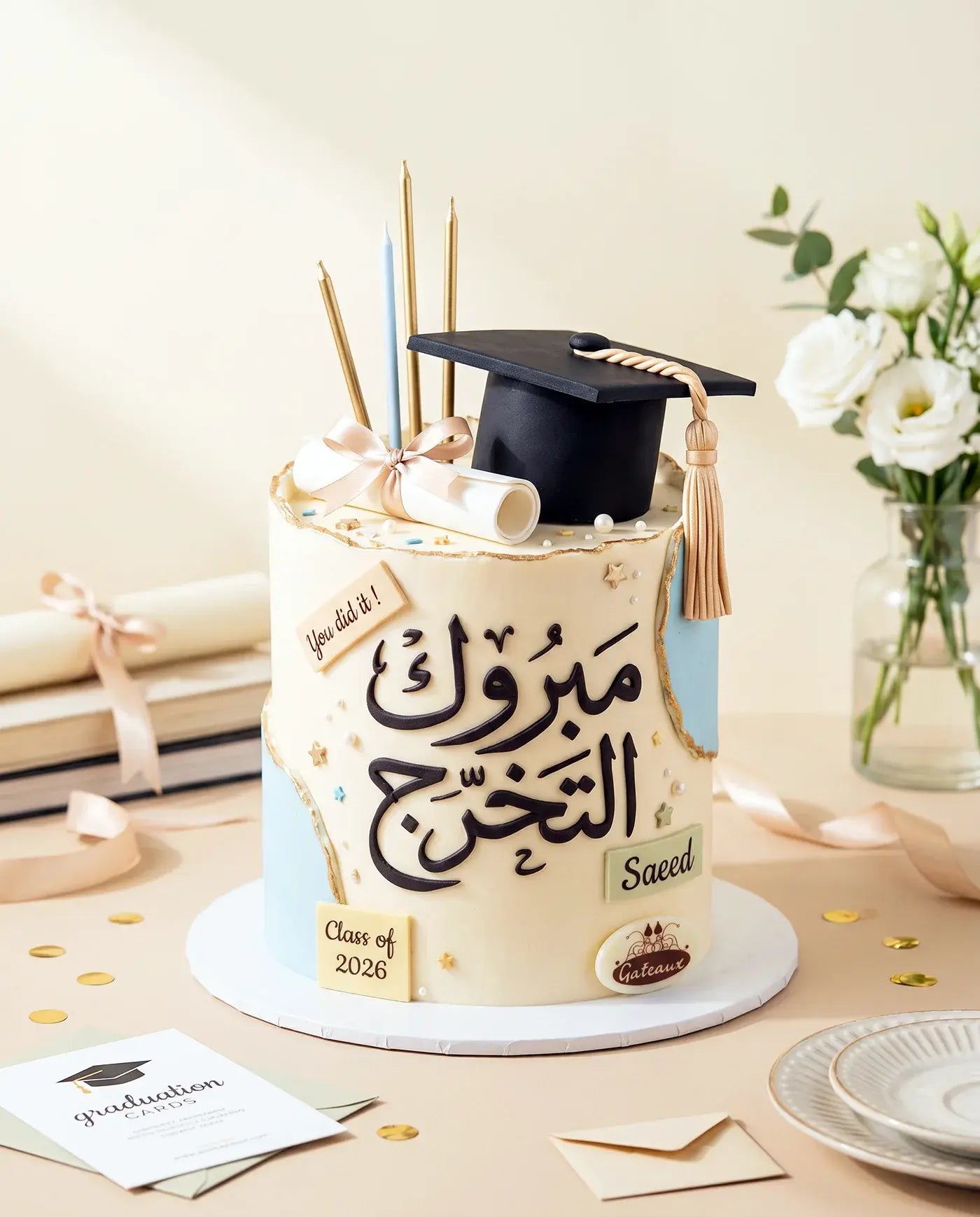 Graduation-themed cake with a black cap and tassel on a table with flowers and stationery.