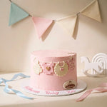 A pink baby shower cake decorated with edible bibs, a pacifier clip, and a heart, displayed on a white board with 'Happy Baby Mom' written in icing. The scene is decorated with pastel bunting and a white rocking horse.