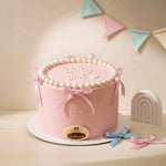 A pink frosted baby shower cake on a white round board, decorated with pearl-like beads around the top edge, pink and blue ribbon bows, and the words 'Baby girl' in white icing. A small oval plaque on the front reads 'Gastone'.