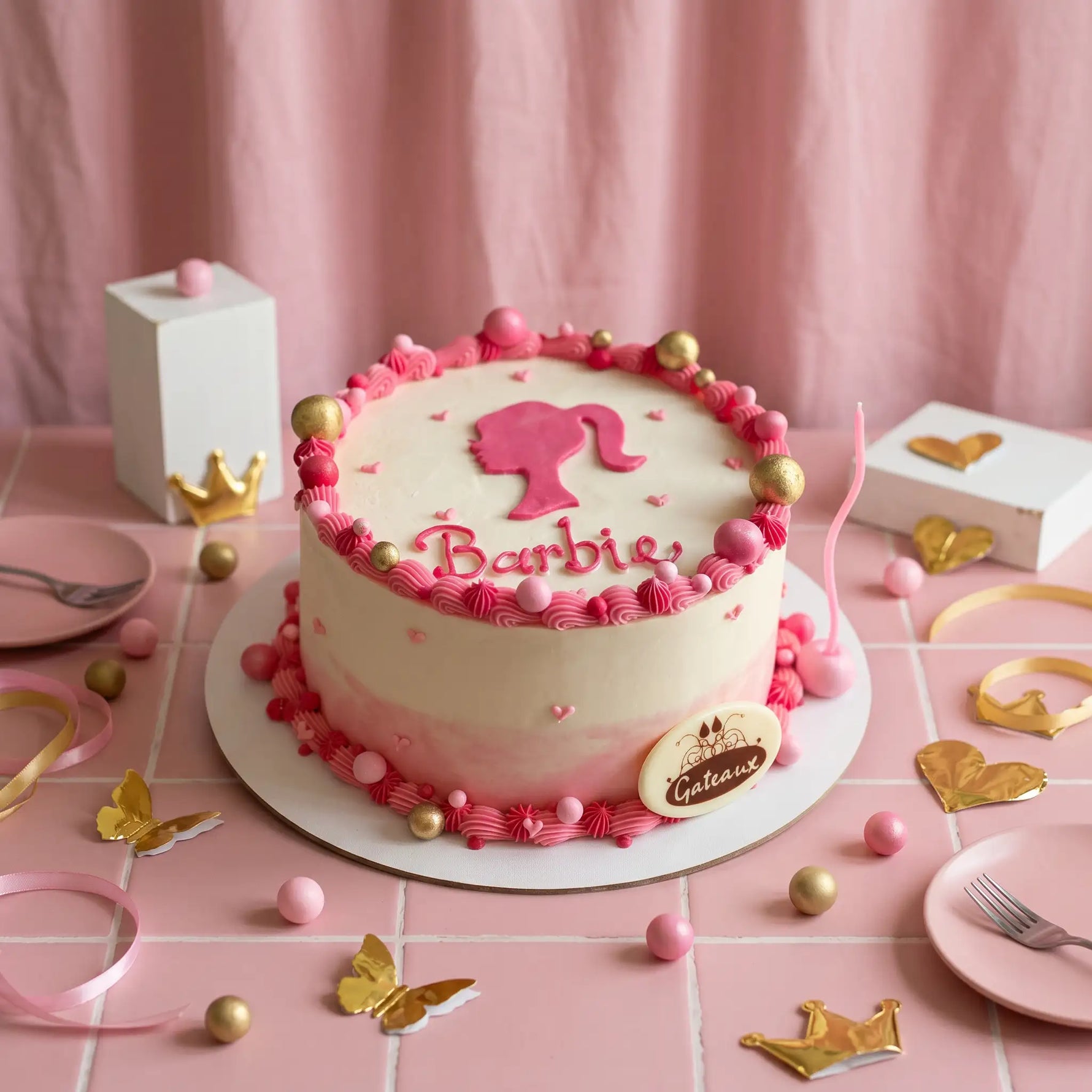 A pink and cream Barbie-themed cake decorated with a pink silhouette of Barbie, the name 'Barbie' in script, and gold and pink accents, displayed on a pink tablecloth with party decorations.