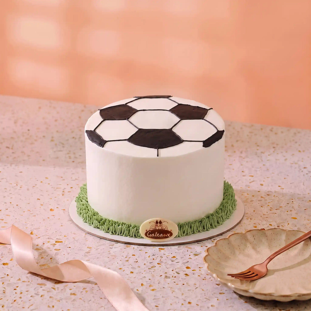A soccer-themed cake with a white frosted body, black and white paneled top, and green grass-like frosting around the base, displayed on a speckled countertop with a peach background.