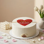 A round white frosted cake on a white plate, decorated with a red heart-shaped topper that reads 'Love 22.02.