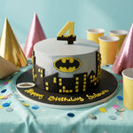 A Batman-themed birthday cake with a white top, black and yellow cityscape base, a yellow number 4 topper, and 'Happy Birthday Schumacher' written on the front. The cake is styled on a light blue tablecloth with party hats and cups in the background.