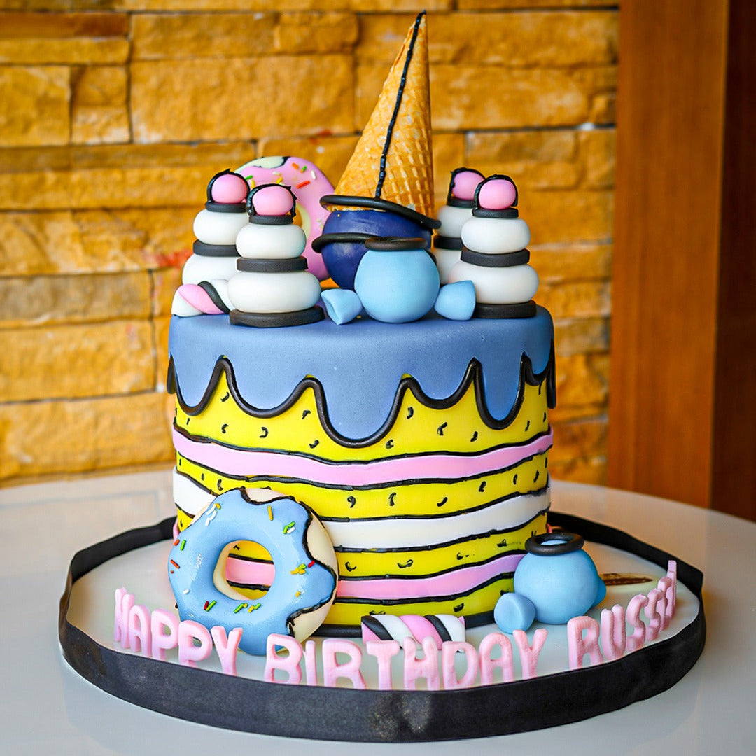 A colorful birthday cake decorated with donut-like shapes and topped with character figures, presented on a table with a 'Happy Birthday' message.