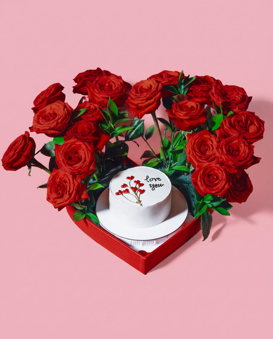 Heart-shaped arrangement of red roses with a small white cake in the center on a pink background