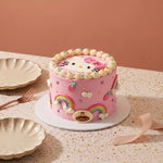 A pink Hello Kitty mini cake decorated with rainbow fondant, white frosting swirls, strawberries, and a Hello Kitty face on top, displayed on a speckled beige table with plates and a ribbon.