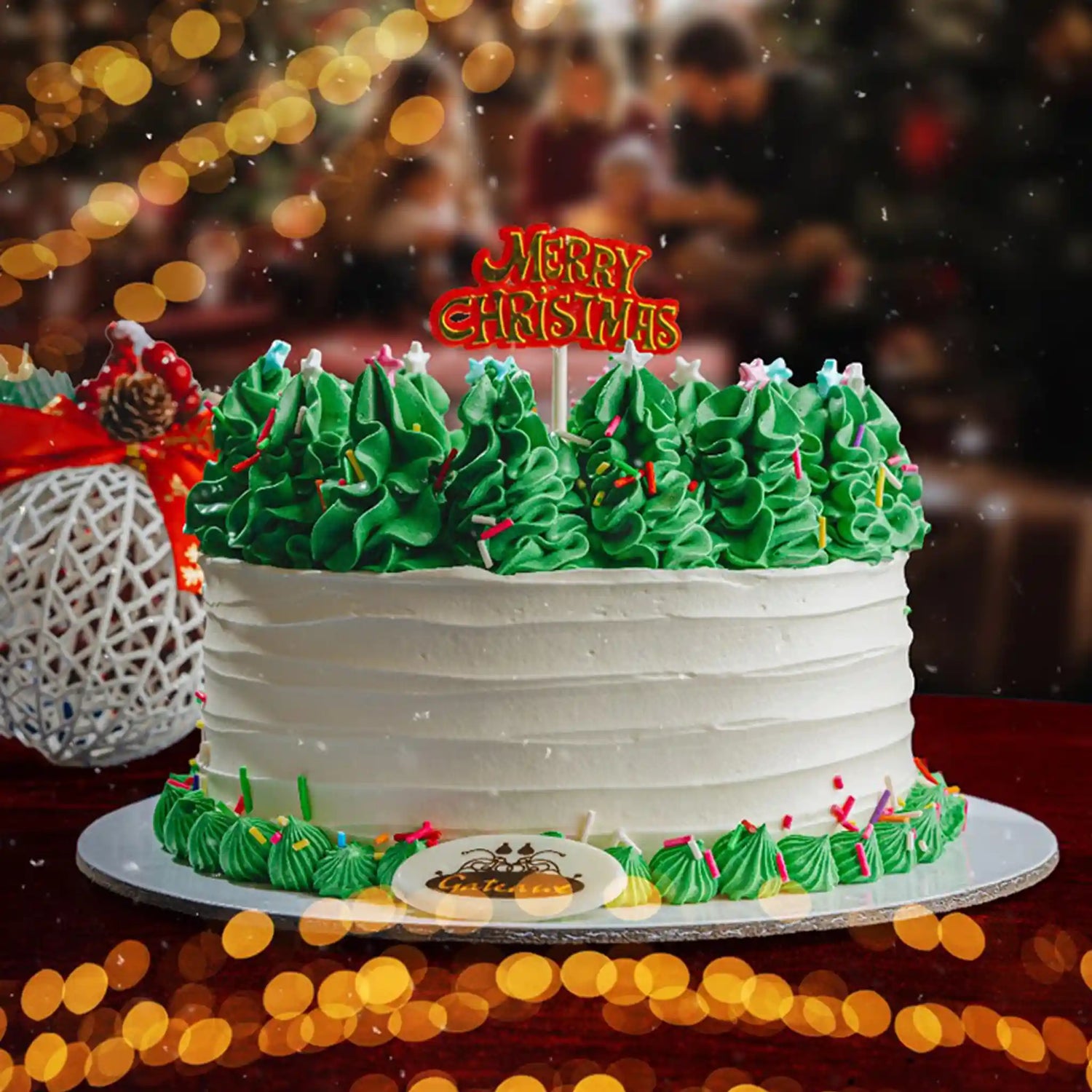 Christmas-themed cake with green trees and 'Merry Christmas' topper, blurred people in the background