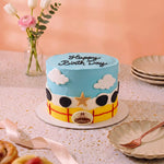 A decorated birthday cake shaped like a cartoon car, with blue frosting sky, white clouds, black wheels, a yellow body, and 'Happy Birthday' written on top, displayed on a table with plates and flowers in the background.
