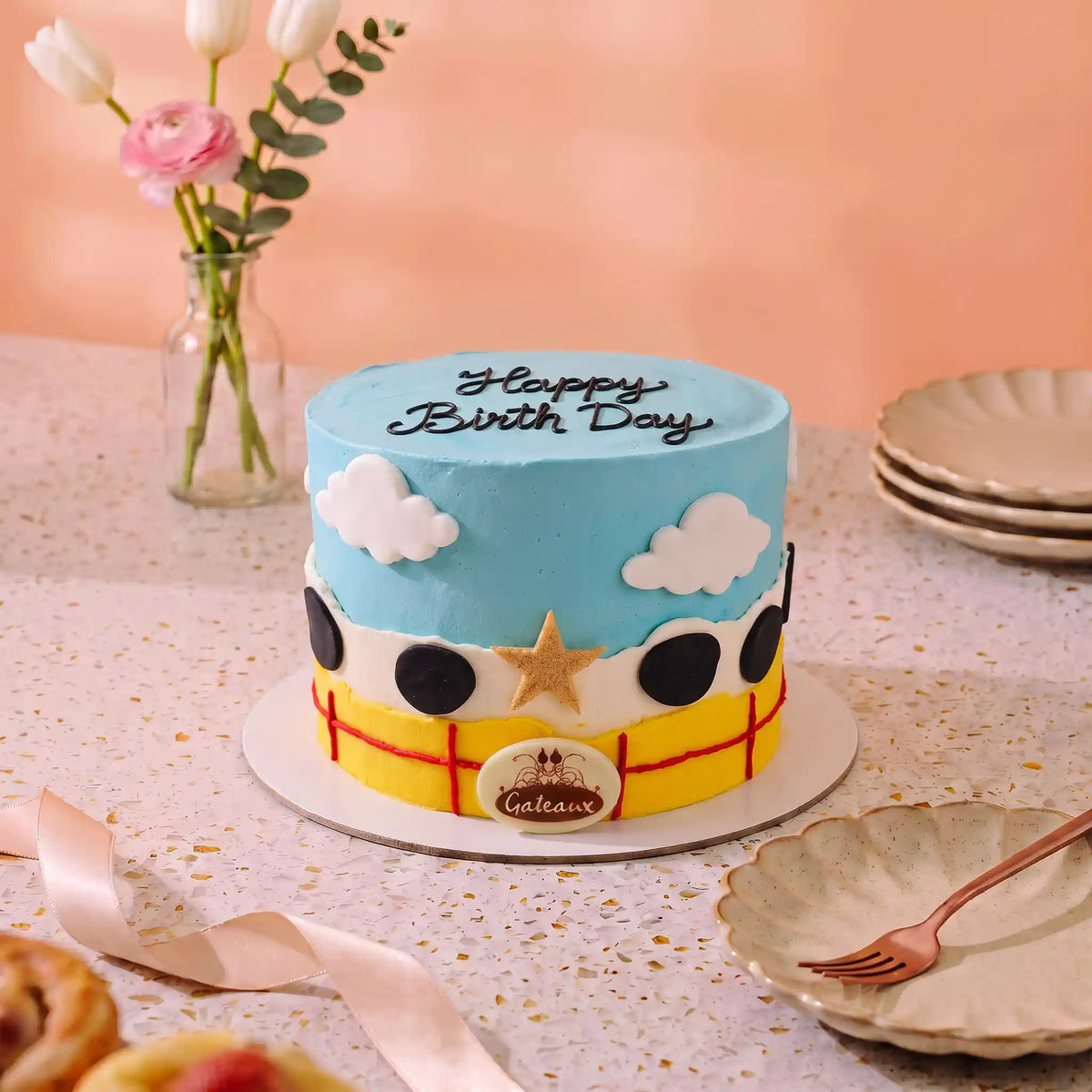 A decorated birthday cake shaped like a cartoon car, with blue frosting sky, white clouds, black wheels, a yellow body, and 'Happy Birthday' written on top, displayed on a table with plates and flowers in the background.