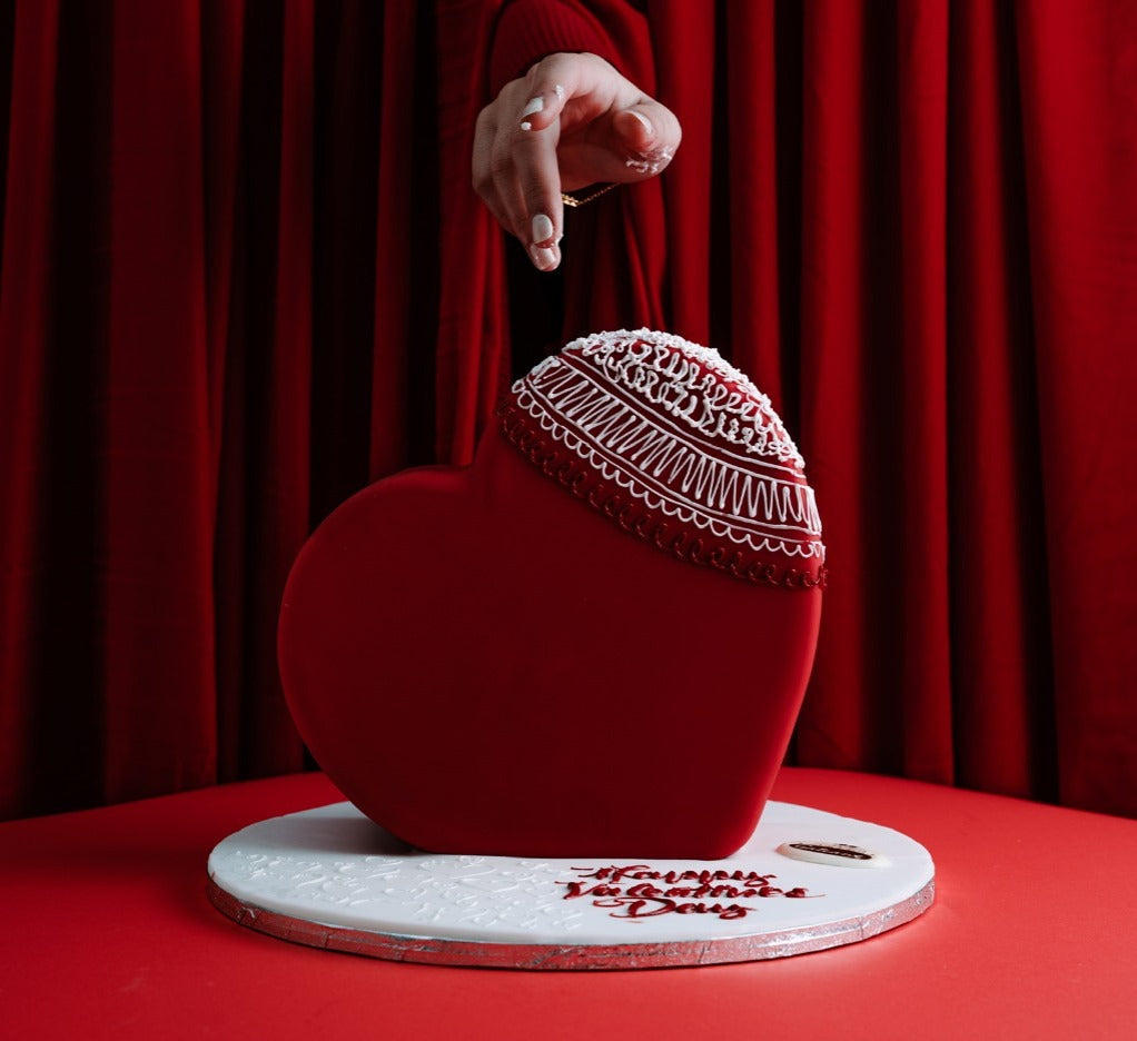 A heart-shaped cake with a white and red design, decorated with a person's hand in the background against a red curtain background.