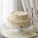A two-tier cream-colored cake with intricate piped floral and lace decorations, displayed on a white cake stand on a round table with plates, baby's breath flowers, and a silver candelabra against a soft white curtain background.