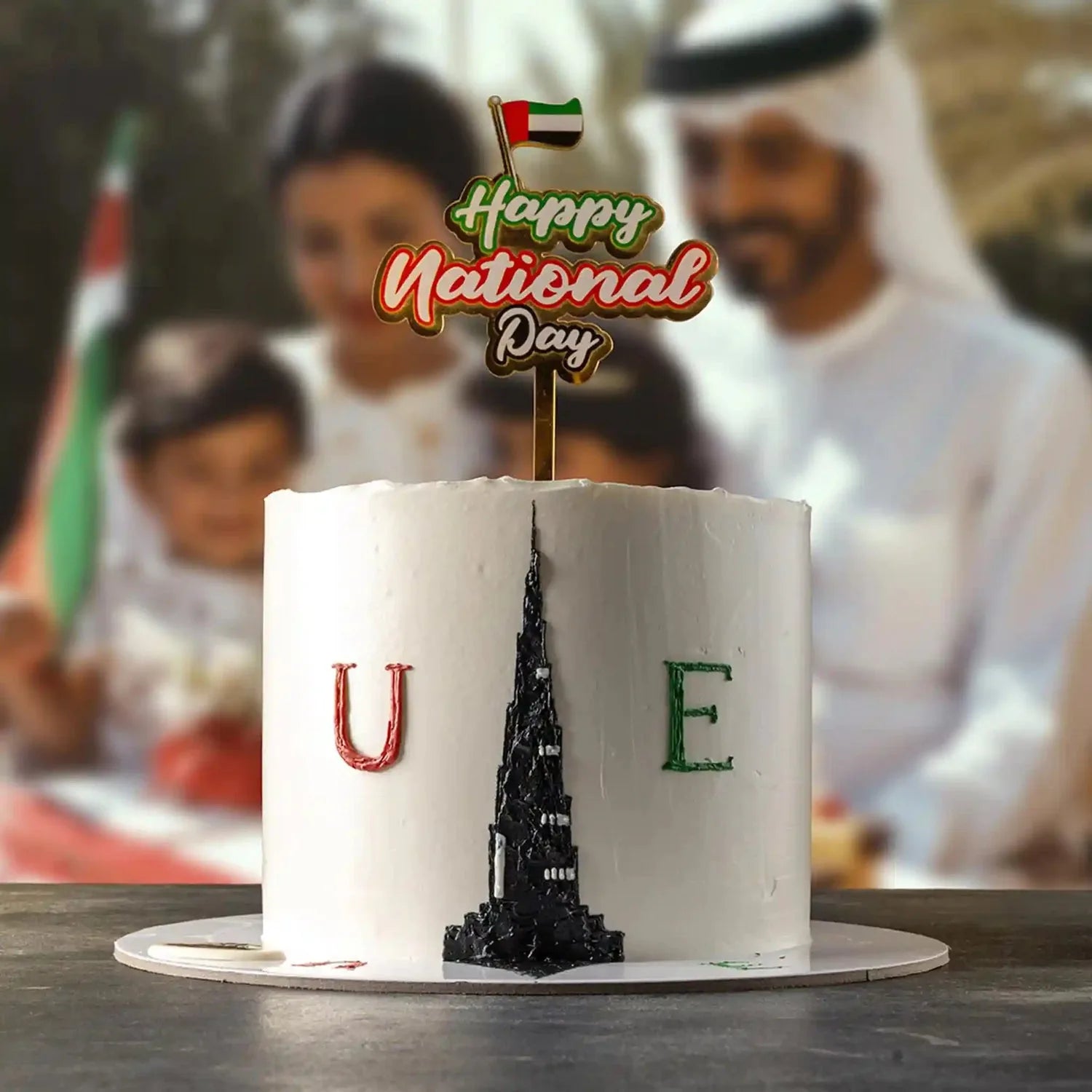 Cake with 'Happy National Day' topper and UAE flag, blurred background of people and flags.