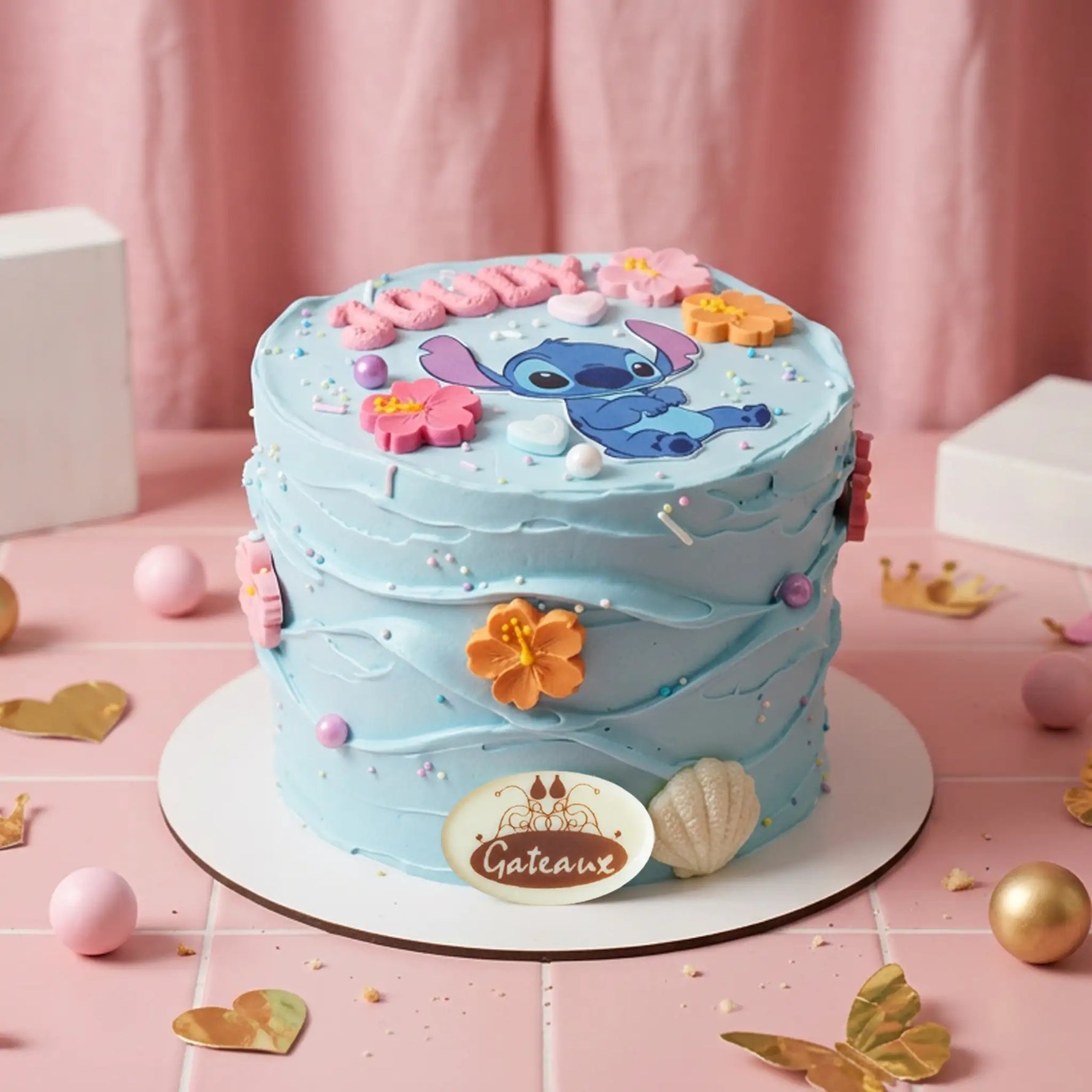 A round light blue frosted cake decorated with a blue cartoon character, pink and orange flowers, and a pink 'Happy Birthday' topper, displayed on a pink surface with decorative props.