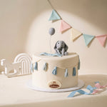 A round white baby shower cake decorated with a gray elephant figurine holding a balloon, blue tassel garlands, and a small plaque on the front, styled on a cream background with pastel bunting and party props.