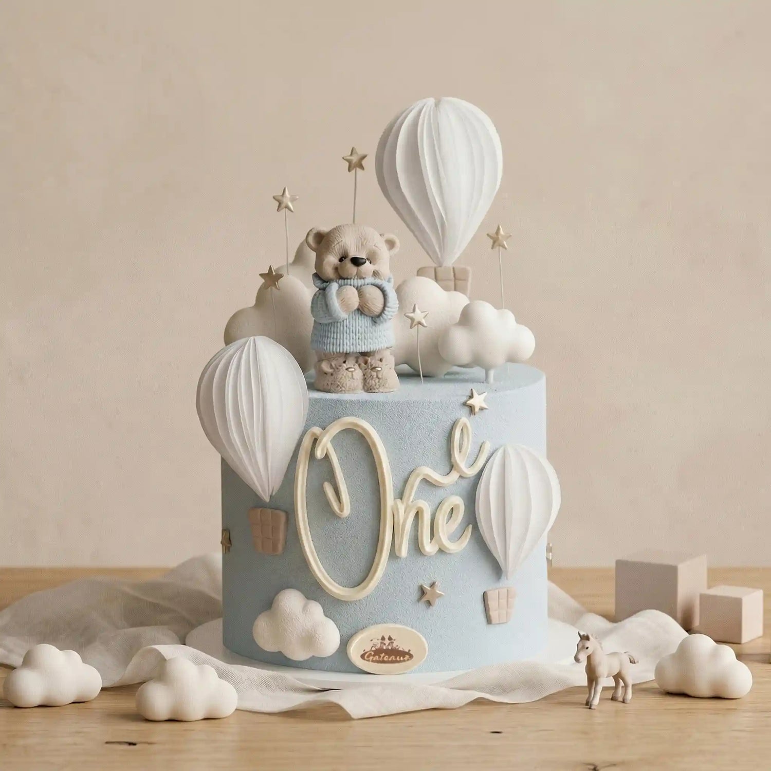 Decorative cake with teddy bear, hot air balloons, and 'One' lettering on a neutral background