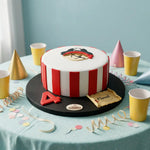 A round birthday cake decorated with red and white vertical stripes and a pirate face topper, displayed on a black round stand on a light blue tablecloth with party hats, cups, and confetti.