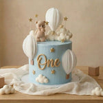Children's birthday cake with teddy bear, hot air balloons, and 'One' decoration on a beige background.