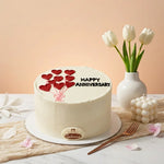 A round white frosted anniversary cake decorated with red heart balloons and the words 'HAPPY ANNIVERSARY' in black icing, displayed on a marble surface with white tulips in a vase and a small white candle nearby.