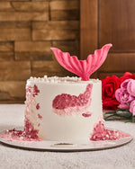 Mermaid-themed cake with pink tail and fish design on a wooden surface with flowers.