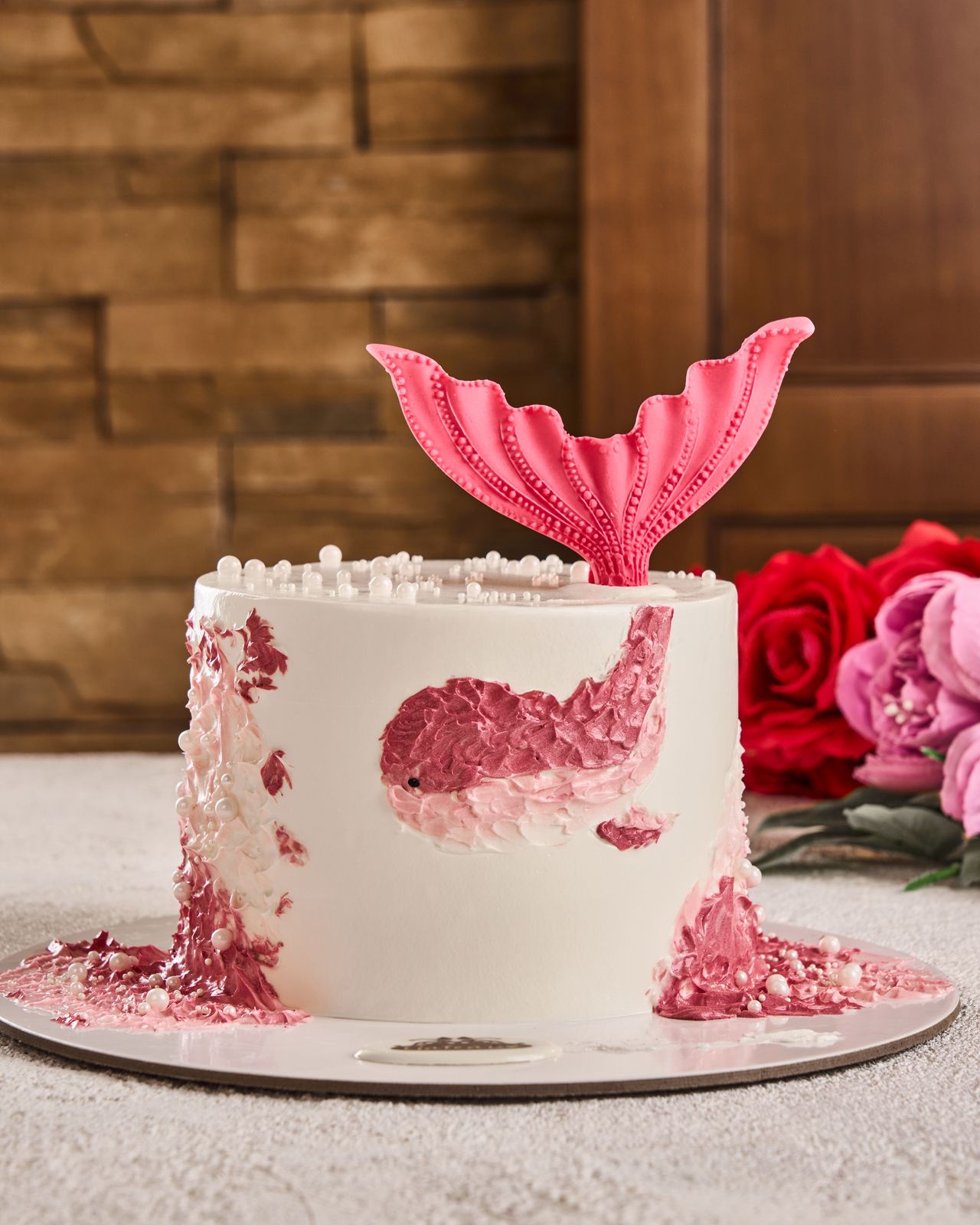 Mermaid-themed cake with pink tail and fish design on a wooden surface with flowers.