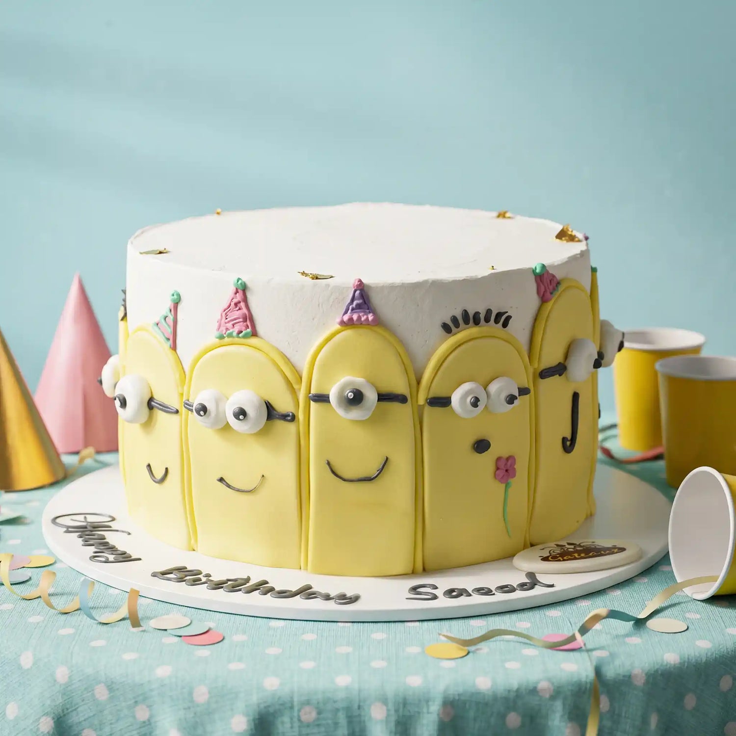 A round birthday cake decorated to look like five yellow cartoon minions with white frosting, party hats, and custom names 'Sasaad' and 'Happy Birthday Sasaad' written on the base, set on a polka-dotted tablecloth with party hats and cups.