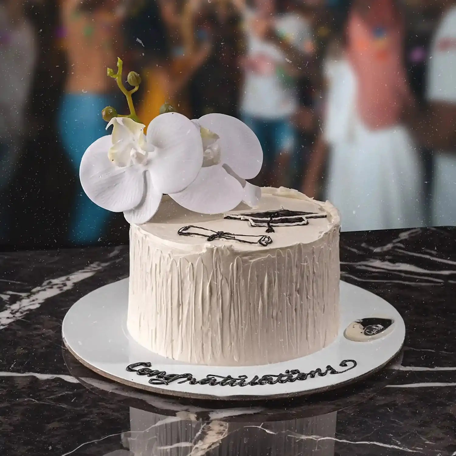 Decorative cake with white flowers on a marble surface, with a blurred background of people.