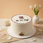 A white frosted Anniversary Cakes
cake with red heart accents and the text 'July 17 It's our day!' on top, displayed on a white cake board. The cake is decorated with a small round 'Galante' logo plaque at the base and sits on a marble surface with tulips in a vase and pink petals around it.
