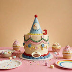 A decorated cake for kids with a blue party hat topper and a red number 3 candle, styled on a pink table with cupcakes, pastel plates, and confetti against a soft peach background.