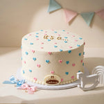 A round Gender Reveal cake with white frosting, decorated with small pink and blue hearts and gold 'it's a' lettering. A small oval plaque on the front reads 'Galante'.