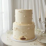 A two-tier cream-colored frosted cake decorated with pearl-like beads, delicate lace trim, and a round chocolate plaque reading 'Galette' on a white round table.