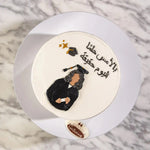 Decorative cake with a figurine of a person in traditional attire and Arabic text on a marble background.