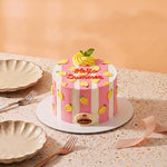 A round mini cake decorated with pink and white vertical stripes, yellow lemon illustrations, and a yellow frosting swirl on top with a green mint leaf. The cake features the text 'Hello Summer' in red icing and a round gold 'Gourmet' label on the front.
