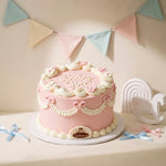A round pink Baby Girl Shower Cake decorated with white piped frosting borders, small pink flowers, bows, and the words 'Baby Girl' on top, displayed on a white board in a styled room setting with pastel bunting and baby-themed decor.