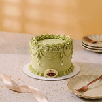 A round green cake with ornate piped frosting decorations and a small round logo plaque reading 'Cielés', displayed on a speckled tabletop with plates and a ribbon in a softly lit setting.