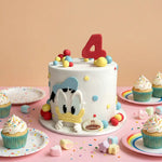 A decorated white cake with a large red number 4 topper and a Donald Duck character face on the front, styled on a pink table with cupcakes, pastel candies, and confetti against a peach background.