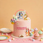 A pink frosted cake decorated with a rainbow topper featuring a dog character, a gold number 2, white daisy flowers, pastel balloon clusters, and the name 'JOUDY' in pink letters. The cake is styled on a pink surface with cupcakes, paper plates, and candy against a peach background.